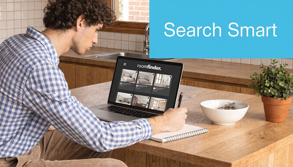 A man wearing a blue plaid shirt using a laptop to search for rooms online at a table.